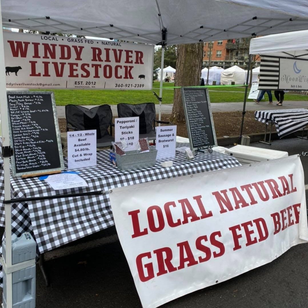 163567923_2935173563475712_1693192405654780656_n Farmers market booth for Windy River Livestock featuring local, natural, grass-fed beef products. The display includes pricing for whole beef cuts, pepperoni, teriyaki sticks, summer sausage, and bratwurst. A banner emphasizes "Local Natural Grass Fed Beef."