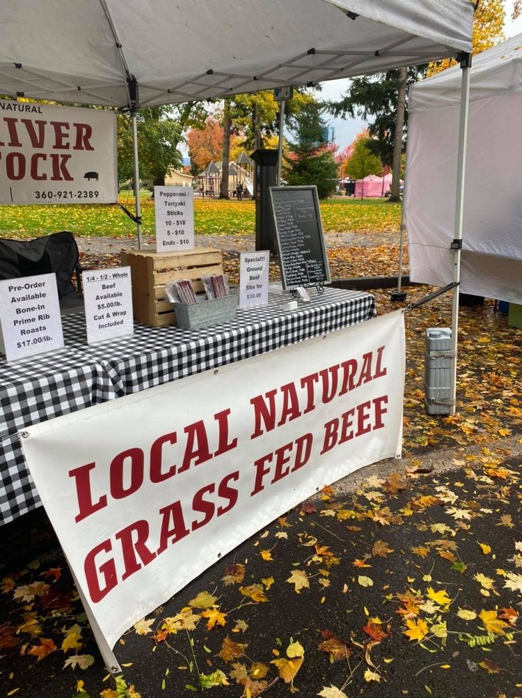 Farmers market stall featuring a banner for local natural grass-fed beef. The display includes prices for various beef products, such as whole beef, ground beef, and pepperoni sticks. A blackboard lists additional offerings and contact information for pre-orders. The setting is surrounded by autumn foliage.