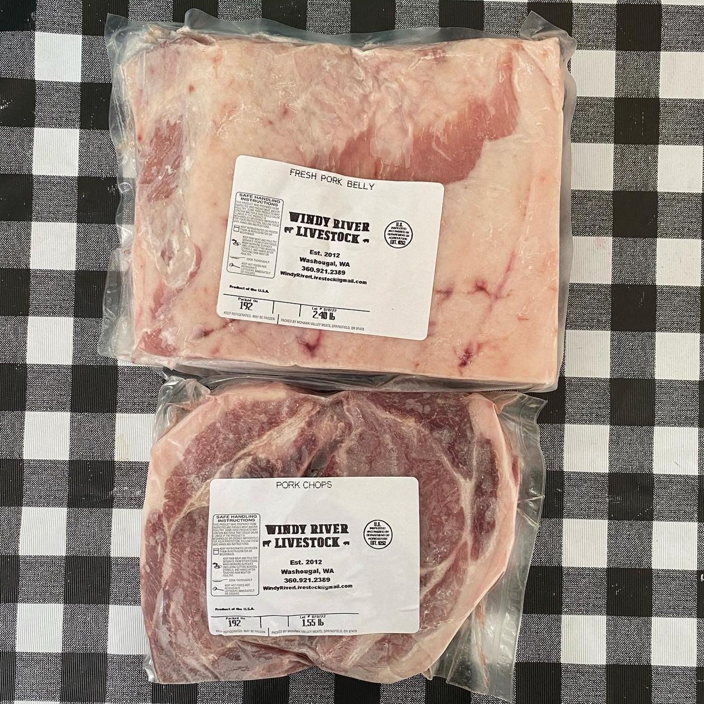 Fresh pork products from Windy River Livestock, including pork belly and pork chops, displayed on a black and white checkered tablecloth. Packaging features product labels with details about the farm, established in 2012 in Washougal, WA.