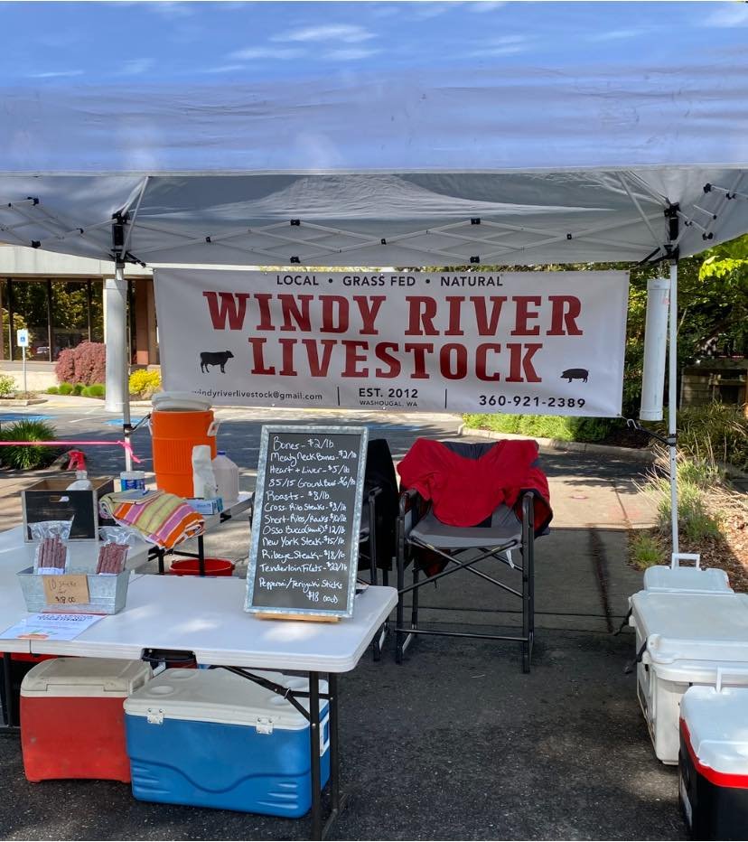 Farmers market booth for Windy River Livestock, featuring a banner with the brand name, contact information, and offerings of local, grass-fed, natural meats. The display includes a chalkboard menu listing various meat prices and types, with coolers and seating visible in the background.