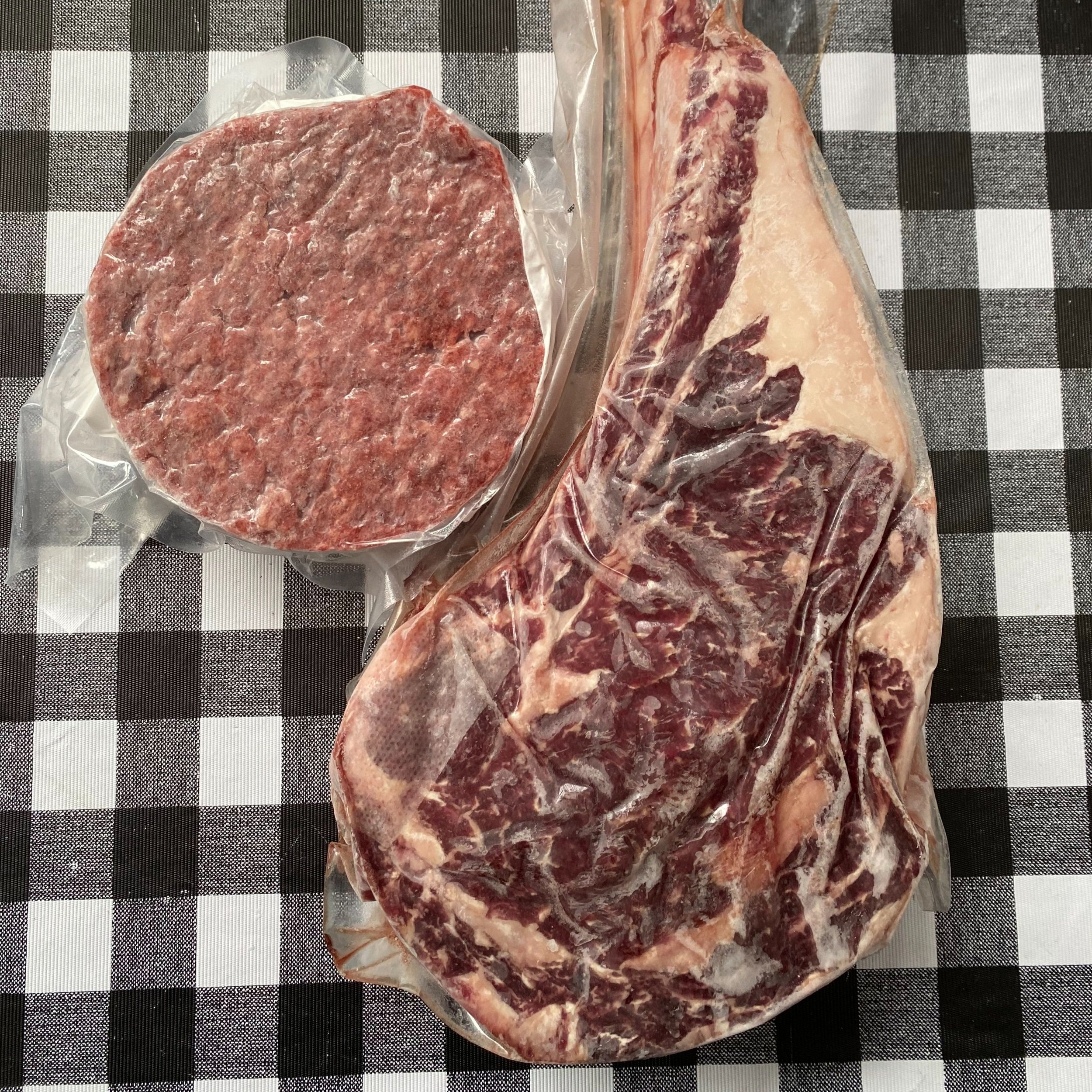 Fresh cuts of beef including a ribeye steak and a hamburger patty, both vacuum-sealed, displayed on a black and white checkered tablecloth.