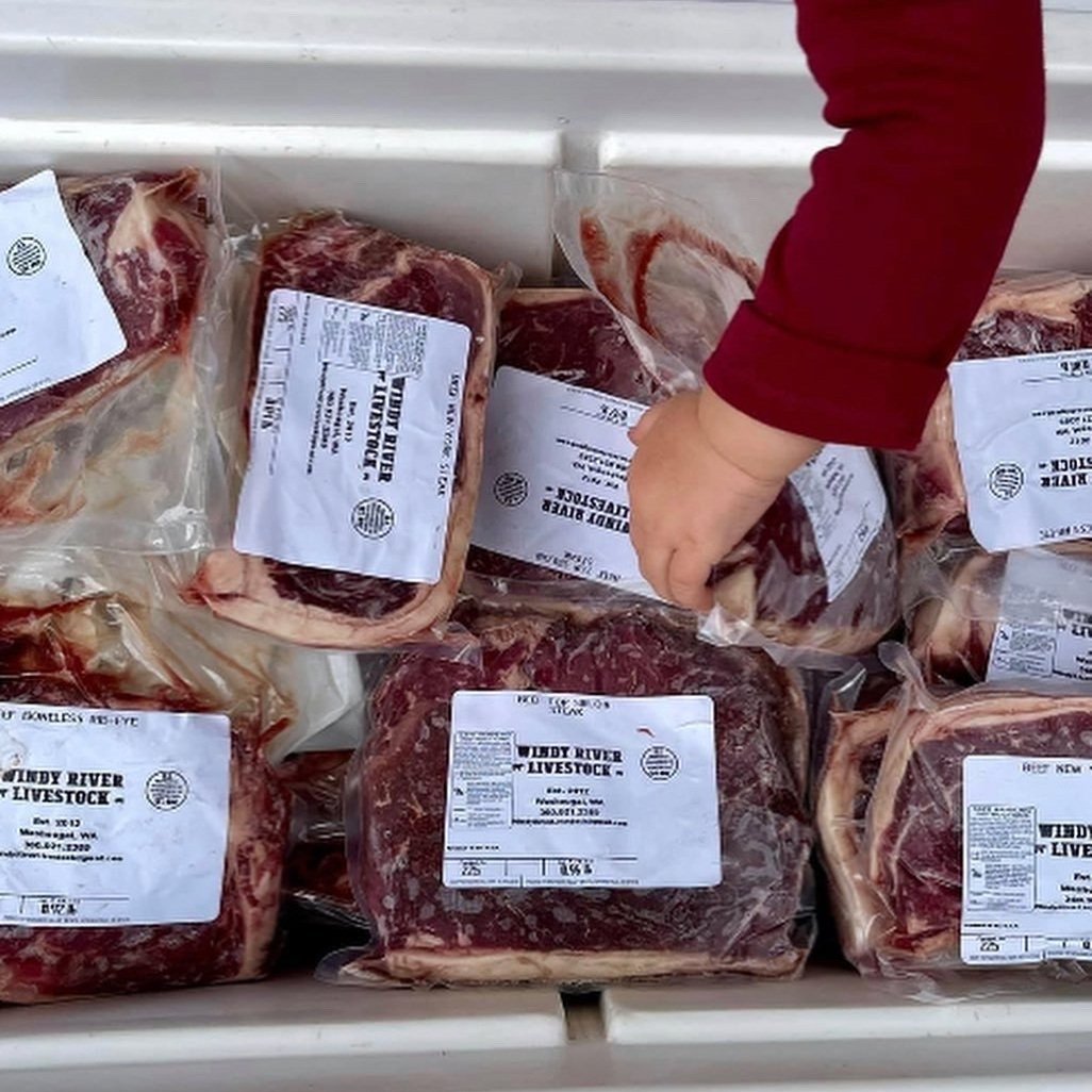 Windy+River+Livestock+-+E90EE989-3DBA-40ED-A11D-97EE1E9C9CFB Packages of beef cuts from Lindy River Livestock displayed in a cooler, with a hand reaching for a rib-eye steak. Each package is labeled with product information and the farm's branding.